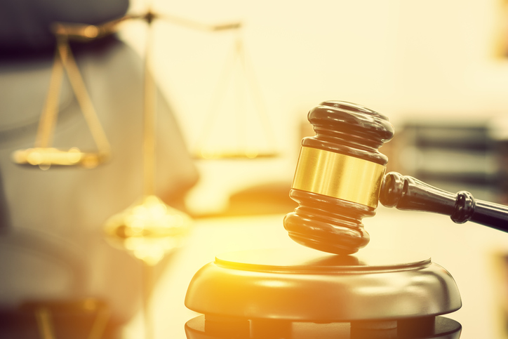 Wooden judge gavel or a wood hammer and a soundboard used by a judge person on a desk in a courtroom with a blurred brass scale of justice behind.