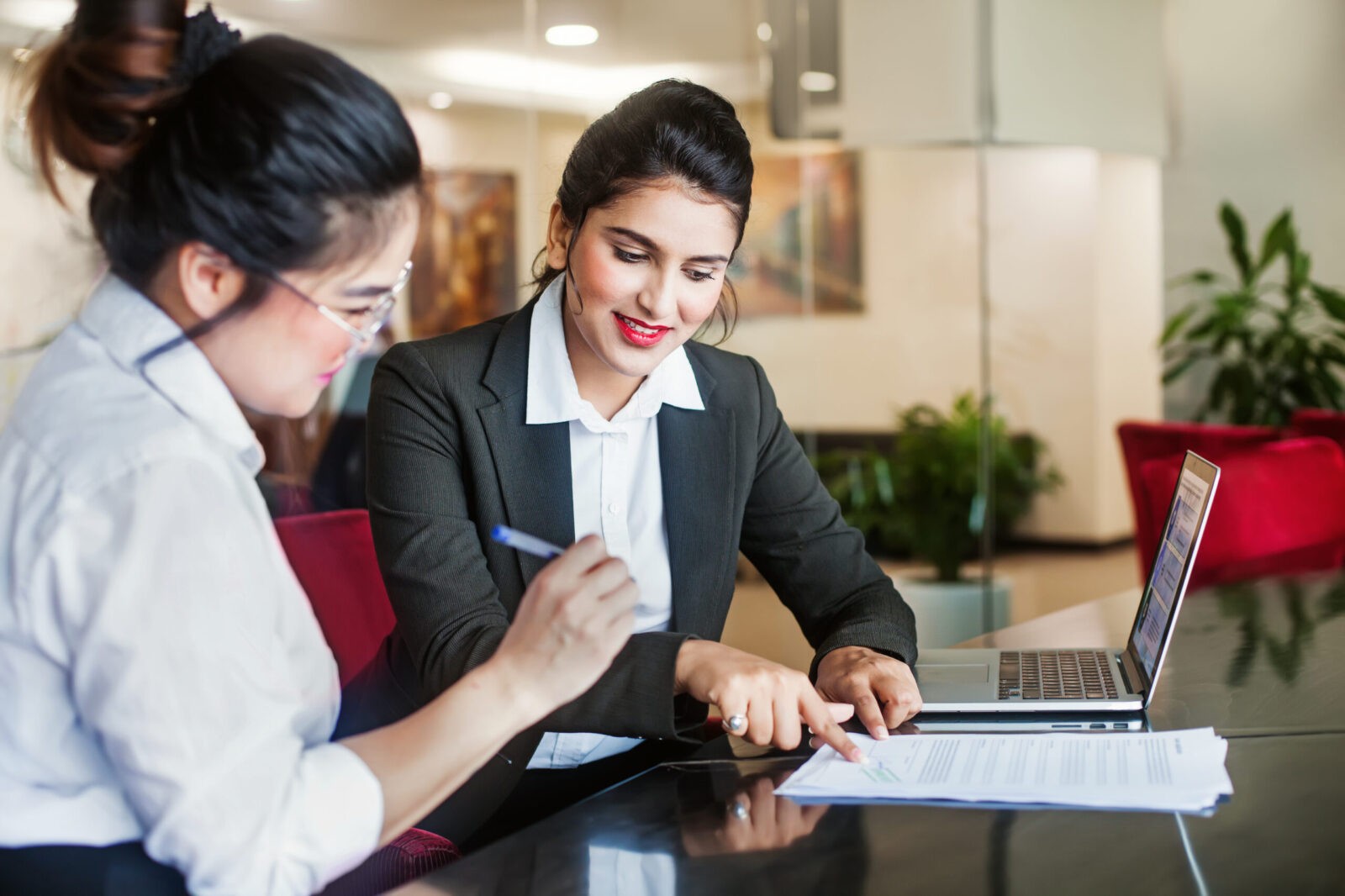 Two women sitting at a desk, one helping the other sign a contract