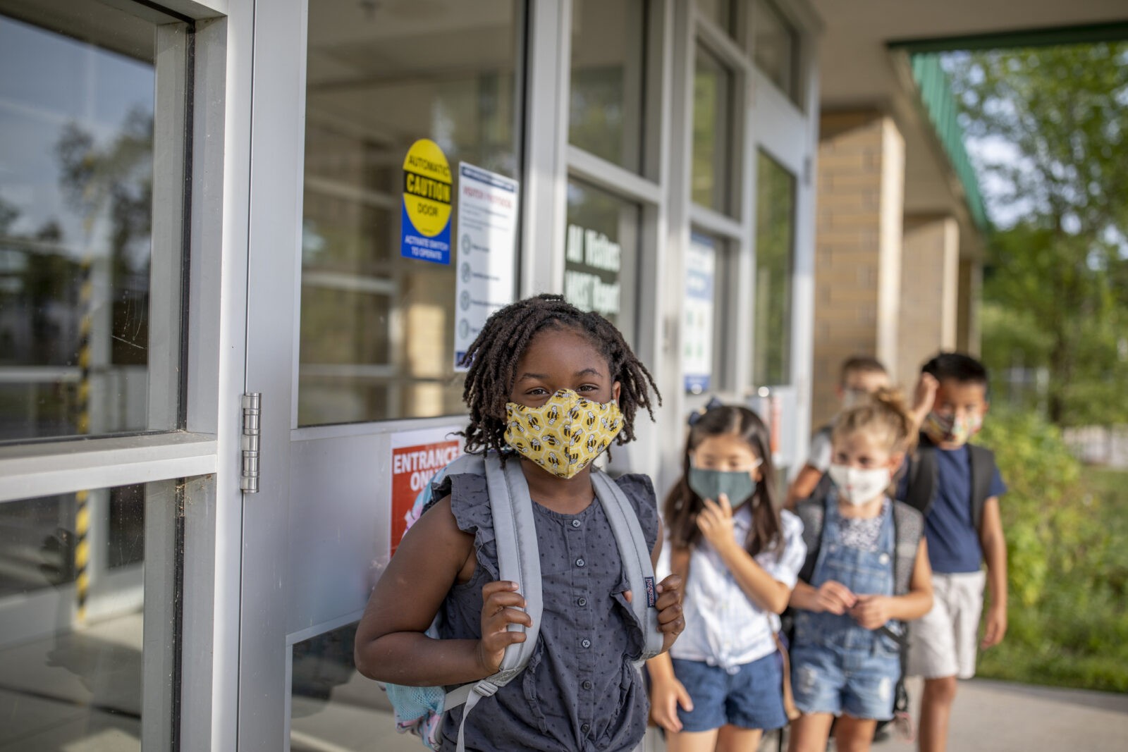 group of little kids in front of a school with backpacks and masks