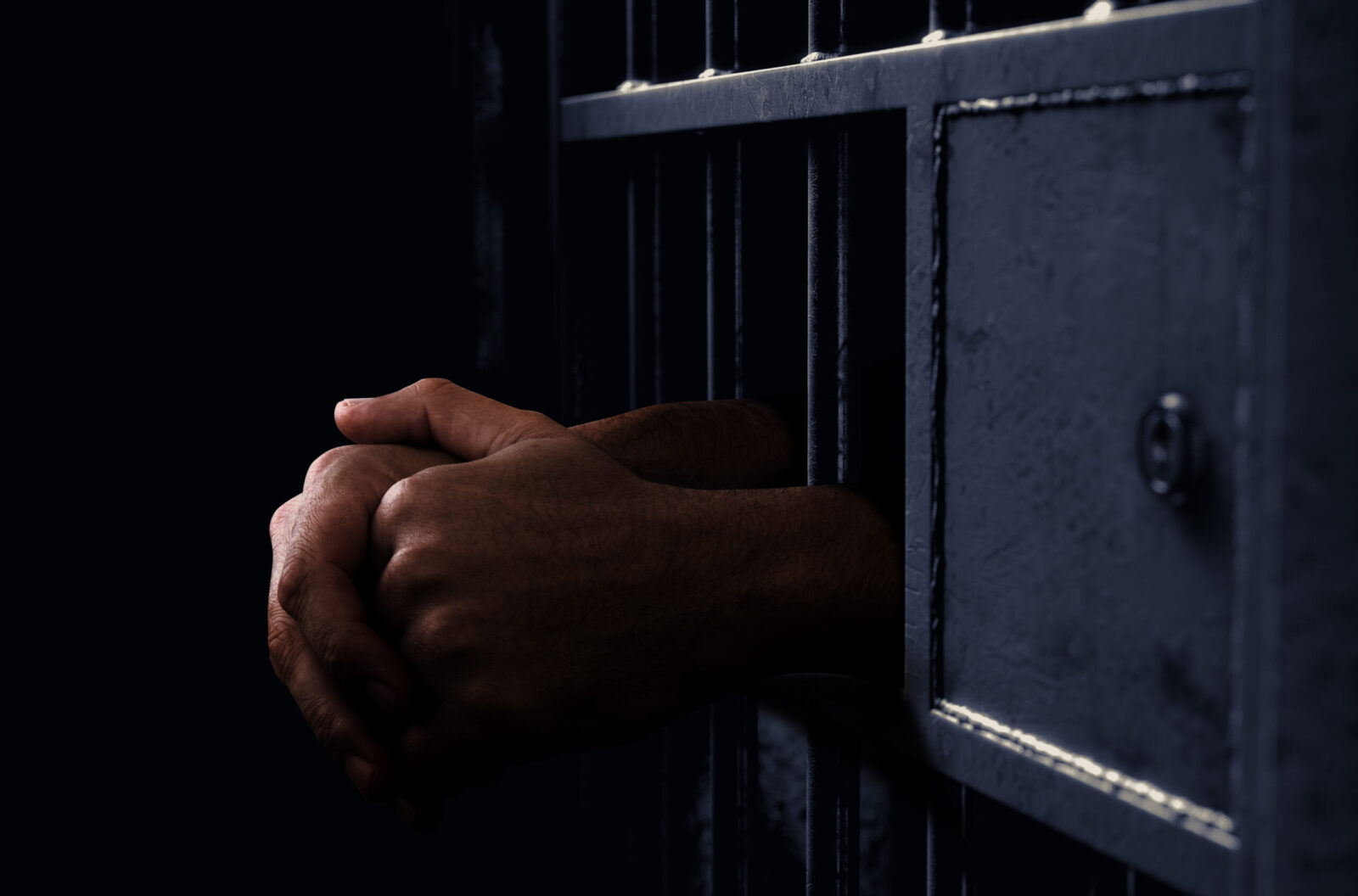 A closeup of a dimly lit prison holding cell door with a pair of hands reaching out in a folded position