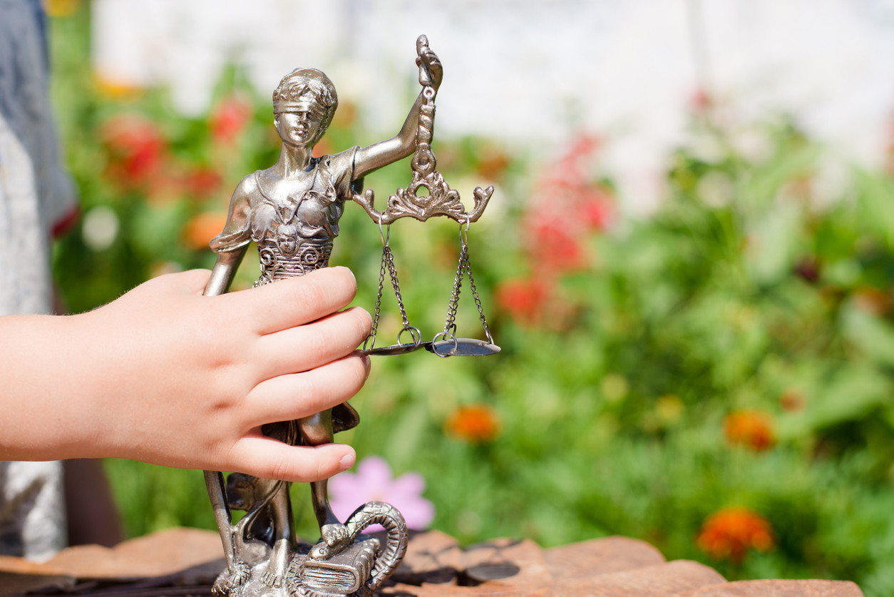 Child's hand holds a bronze statue of Lady Justice with scales, outdoors in a garden.